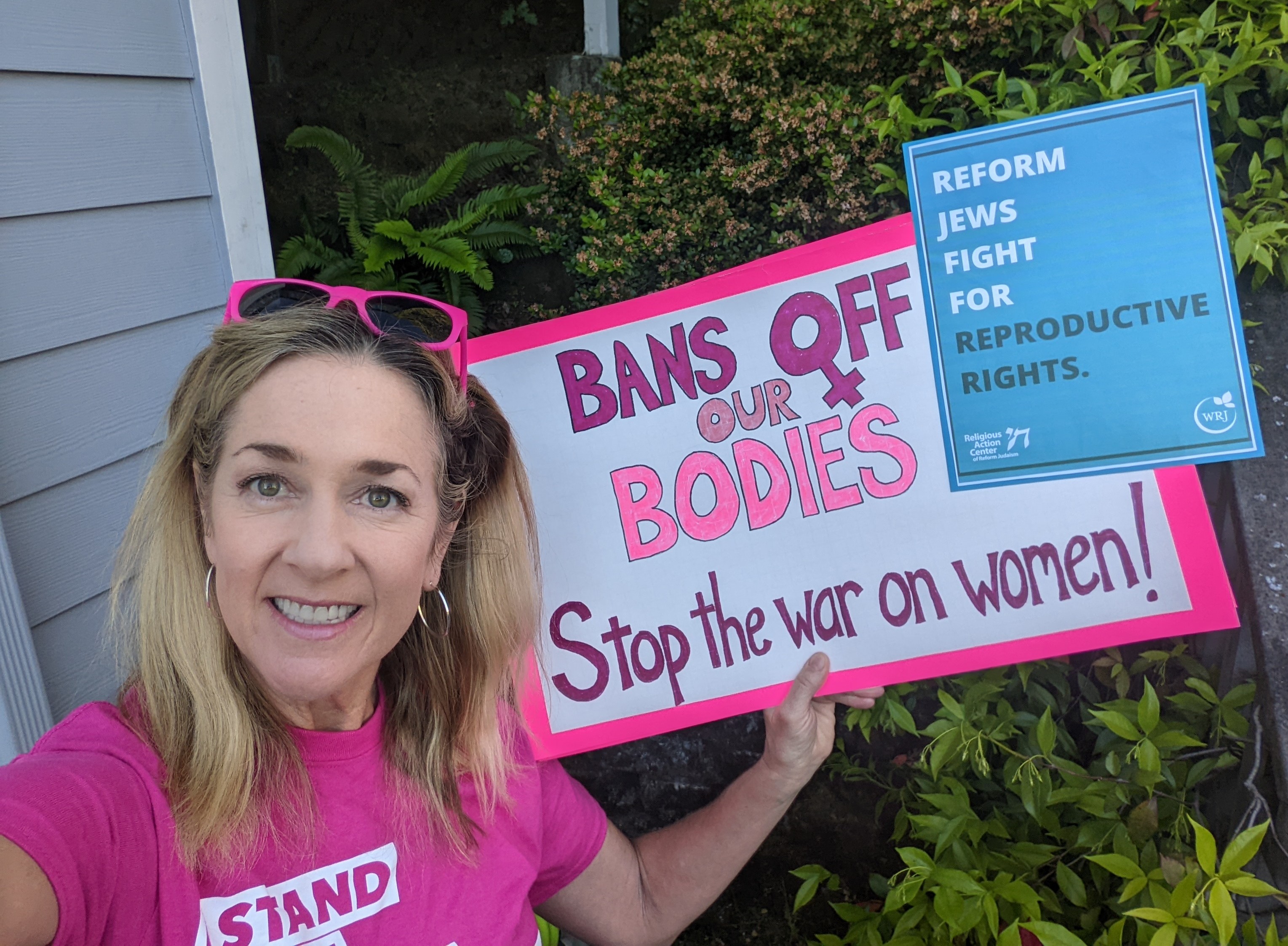 A woman with blond hair and pink sunglasses holds a "Bans off our Bodies Stop the War on Women" banner. 