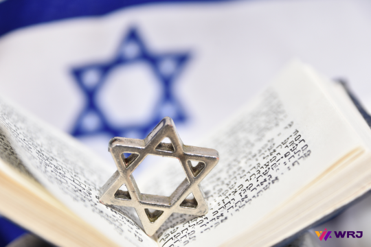 A picture of a half-opened Hebrew book with a silver Star of David placed in the middle. The Israel flag is in the background. 